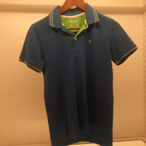 Blå pikétröja från Champion - Säljer en snygg blå pikétröja från Champion med gröna detaljer och broderad logga på bröstet. Tröjan har korta ärmar och en klassisk krage med knappar. Perfekt för en sportig look!