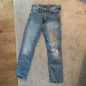 Levi's Jeans - Säljer ett par klassiska blå Levi's jeans med en avslappnad passform. De har en snygg slitning på ena benet och den ikoniska läderpatchen baktill. 