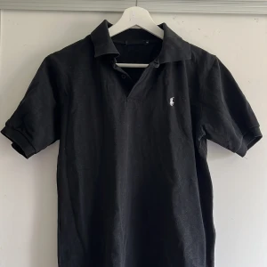 Svart pikétröja från Ralph Lauren - Snygg svart pikétröja från Ralph Lauren. Tröjan har en liten broderad logotyp på bröstet och korta ärmar. Perfekt för en stilren look. 