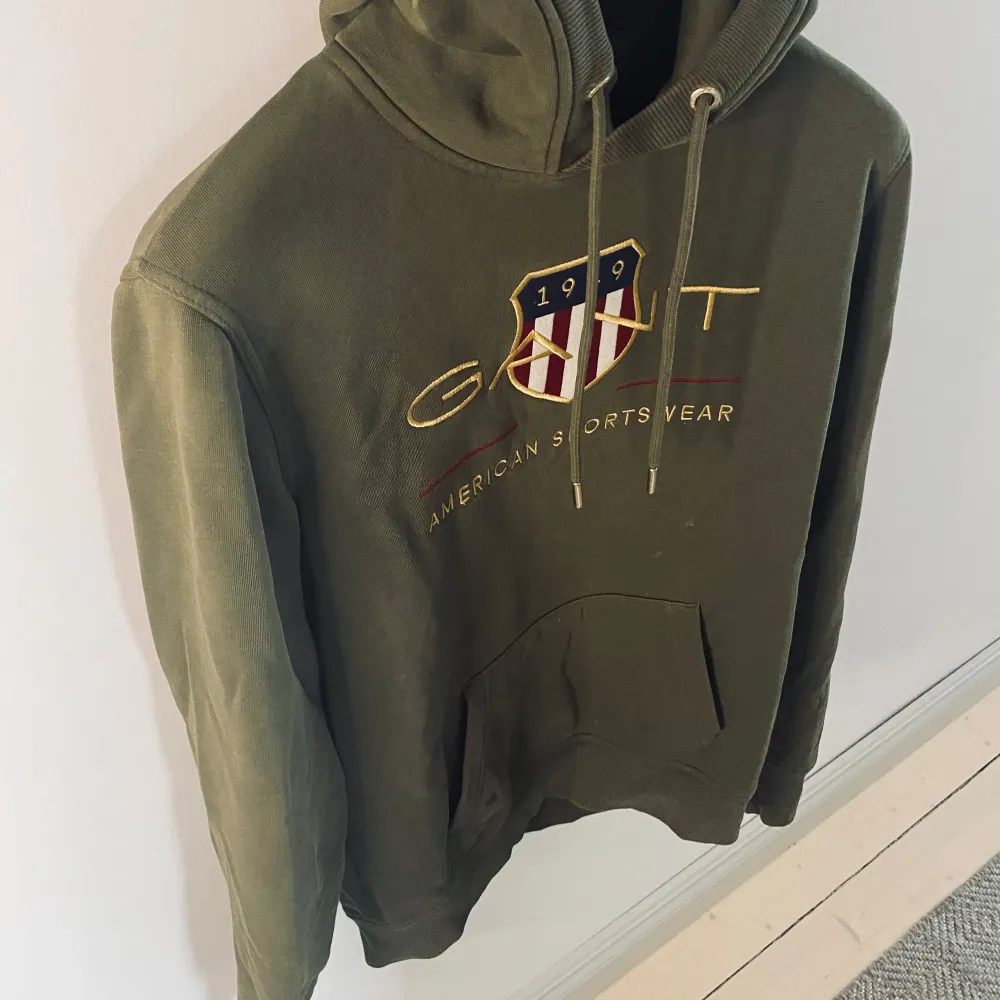 Grön hoodie från GANT med broderad logga.. Hoodien har en klassisk känguruficka och justerbar dragsko i huvan. Så snygg!. Neuletakit & Villapaidat.