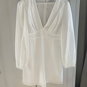 Vit klänning med v-ringning - Superfin vit klänning med djup v-ringning och långa ärmar. Perfekt för en stilren look. Klänningen har en enkel design med en diskret dragkedja i ryggen. Passar perfekt till sommaren och studenten