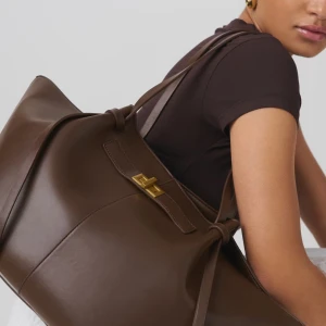 Ginatricot - Long strap pu bag - Snygg brun axelväska i skinn med elegant guldfärgad låsdetalj. Väskan har en rymlig design med långa handtag och knytband på sidorna för justerbarhet. Perfekt för att bära med sig allt man behöver med stil. Är helt nytt och oanvändt! Tyckte om den i början men är trött på den nu🙂