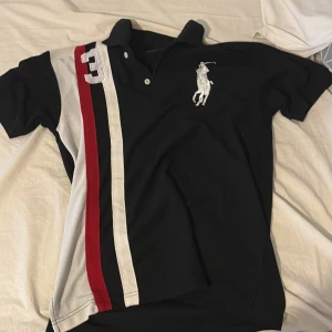 Svart pikétröja från Ralph Lauren - Snygg svart pikétröja från Ralph Lauren med stora broderade detaljer. Tröjan har korta ärmar och en klassisk krage. Färgglada ränder i rött, vitt och grått på framsidan ger en sportig look.