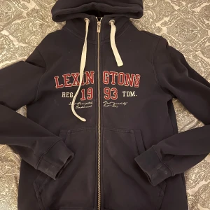 Mörkblå hoodie från Lexington - Säljer en mörkblå hoodie från Lexington med dragkedja och vit text på framsidan. Den har en justerbar huva med vita snören och långa ärmar. Perfekt för en avslappnad stil.
