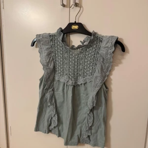 Grön ärmlös blus från Mango - Söt grön ärmlös blus från Mango med spetsdetaljer och volanger längs ärmhålen. Blusen har ett vackert broderat mönster framtill och en rund halsringning. Perfekt för en somrig look!