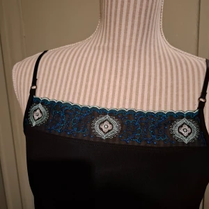 Svart klänning med blå spetsdetaljer - Elegant svart klänning med smala axelband och vackra blå spetsdetaljer vid halsringning och nederkant. Klänningen har en figurnära passform och når till knäna. Perfekt för en stilfull kväll.