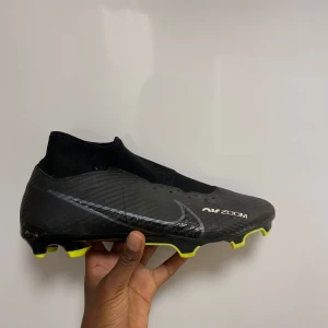 Svarta Nike fotbollsskor med gula detaljer - Snygga svarta Nike fotbollsskor med gula detaljer och en strumpliknande passform. Skorna har en modern design med Nike Zoom-teknologi för extra komfort och prestanda på planen.