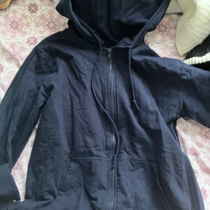 Svart huvtröja från Tezenis - Säljer en svart huvtröja från Tezenis med dragkedja och fickor framtill. Tröjan har långa ärmar och en justerbar huva med snören. Perfekt för en avslappnad stil. Den är ny men har aldrig använt den. Köpte den 2022 men den har bara varit i min garderob. Den är dock tunn så den rekommenderas att ha på sig inomhus för gymmet, hemmet eller under en kall sommar dag.