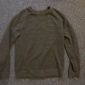 Olivgrön stickad tröja från Ralph Lauren - Säljer en stilren olivgrön stickad tröja från Ralph Lauren. Tröjan har långa ärmar och ribbade muddar vid ärmslut och nederkant. Perfekt för kyligare dagar med sin klassiska design och diskreta logga på bröstet.