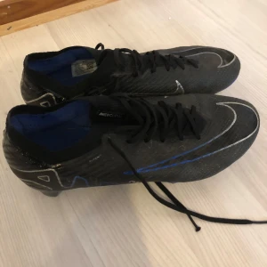 Nike Mercurial Vapor 15 Elite FG - Säljer ett par svarta Nike fotbollsskor med blå och vita detaljer. Skorna har snörning och är designade för optimal prestanda på planen. Perfekta för fotbollsspelare som söker både stil och funktion. Skick på dom här 8,5/10. Priset kan diskuteras vid snabb affär🔥