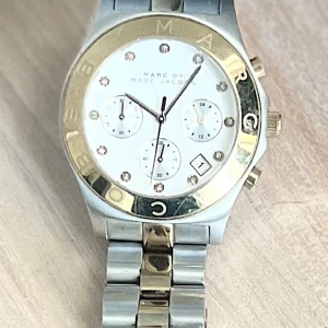 Klocka från Marc by Marc Jacobs - Snygg klocka från Marc by Marc Jacobs med en stilren design. Urtavlan är vit med guldiga detaljer och tre mindre urtavlor. Armbandet är i silver och guld, vilket ger en elegant touch. Perfekt för den som vill ha en tidlös accessoar.