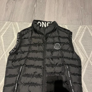 Svart dunväst från Moncler - Snygg svart dunväst från Moncler med dragkedja och logotyp på bröstet. Västen har en quiltad design och hög krage. Perfekt för kyligare dagar.