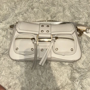 Vit axelväska med kedjedetalj - Snygg vit axelväska från Aldo med guldfärgade kedjedetaljer och två dragkedjefickor framtill. Väskan har en justerbar axelrem och dekorativa knytband. Perfekt för att ge en stilren touch till din outfit.