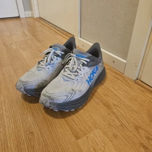 Grå och blå Hoka One One löparskor - Säljer ett par grå och blå Hoka One One löparskor med snörning. Skorna har en robust sula och är designade för komfort och stöd under löpning. Perfekta för den som söker en pålitlig träningssko.
