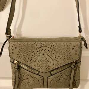 Mossgrön axelväska med perforerat mönster - Snygg mossgrön axelväska med ett unikt perforerat mönster. Köpt i USA. Väskan har två dragkedjor framtill och en justerbar axelrem. Mkt sparsamt använd. Storlek: 25x20