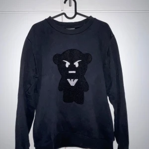 Svart tröja från Emporio Armani med angry taddy som logga - OBS: Pris kan diskuteras samt vid snabb affär kan sänkas pris.                                 Säljer en svart tröja från Emporio Armani med en cool björndetalj på framsidan. Perfekt för en stilren och avslappnad look. Passar bra med gråa eller svarta jeans eller byxor men allmänt alla passar de bra med. 