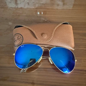 Blå solglasögon från Ray-Ban - Snygga Ray-Ban solglasögon med blå spegelglas och guldfärgad metallram. De klassiska pilotglasögonen kommer med ett brunt fodral för säker förvaring. Perfekta för att ge en cool touch till din stil. Pris kan diskuteras :) 