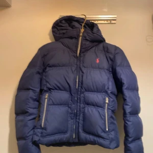 Blå dunjacka från Polo Ralph Lauren - Snygg blå dunjacka från Polo Ralph Lauren med huva och dragkedja. Jackan har två praktiska fickor med dragkedjor och en ikonisk röd logga på bröstet. Perfekt för kyliga dagar.