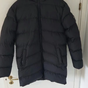 Svart dunjacka med huva - Säljer en svart dunjacka med huva. Jackan har långa ärmar och en dragkedja framtill. Perfekt för kalla dagar.