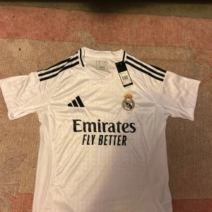 Real Madrid fotbollströja  - Snygg vit Real Madrid fotbollströja från Adidas med korta ärmar och klassiska svarta ränder på axlarna. Tröjan har klubbens emblem på bröstet och sponsorlogga 'Emirates Fly Better' tryckt på framsidan. Pris kan diskuteras!! Tveka inte att skriva!!