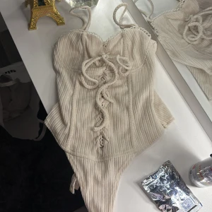 Beige ribbad korsett - Säljer en snygg beige ribbad korsett body med snörning framtill och smala axelband. Den har fina spetsdetaljer vid kanterna. Strl UK 4 motsvarar xs-s 