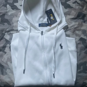 Vit hoodie från Polo Ralph Lauren - Säljer en stilren vit hoodie från Polo Ralph Lauren med dragkedja och den klassiska loggan på bröstet. Storlek L men passar även medium. Perfekt för en avslappnad look. Den har en justerbar huva med snören, pris går att diskutera vid snabb affär.