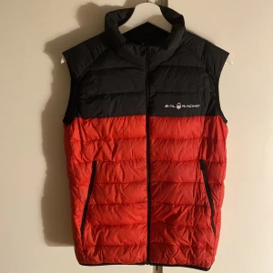 Röd och svart dunväst från Sail Racing i nyskick! - Snygg röd och svart dunväst från Sail Racing med dragkedja framtill. Västen har en quiltad design och två praktiska fickor. Perfekt för kyligare dagar. 10/10 skick!