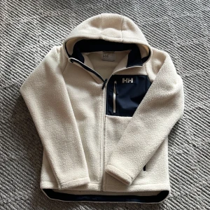 Vit fleecejacka från Helly Hansen - Säljer en vit fleecejacka från Helly Hansen med huva och dragkedja. Jackan har en praktisk bröstficka med dragkedja och är perfekt för kyliga dagar. Den är mjuk och bekväm med en stilren design. Super bra skick! Inga defekter skriva vid frågor och funderingar!
