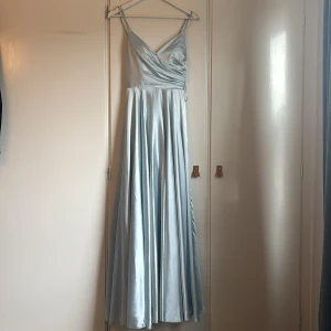 Ljusblå långklänning med snörning - Elegant ljusblå långklänning i satin med omlott-design och smala axelband. Klänningen har en vacker snörning i ryggen som ger en fin detalj och perfekt passform. Perfekt för speciella tillfällen. Storleken är xs men passar jag som är 170 cm lång.