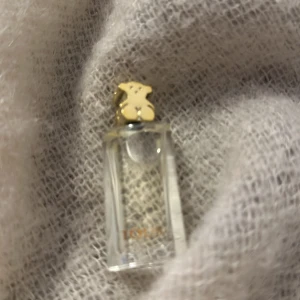 Parfym från Tous - Elegant parfym från Tous i en stilren, rektangulär flaska med en guldig björnformad kork. Förpackningen är i en lyxig guldig färg med präglat mönster. Perfekt för den som älskar en sofistikerad doftupplevelse.