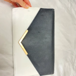Svart och vit kuvertväska med guldkedja - Elegant kuvertväska i svart och vitt med en stilren guldkedja. Väskan har en triangulär design med en snygg gulddetalj på framsidan. Perfekt för att ge en touch av lyx till din outfit.
