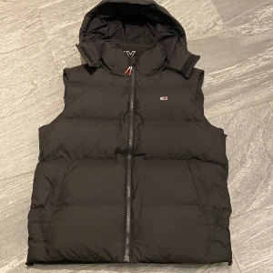 Svart dunväst med avtagbar huva - Snygg svart dunväst från Tommy jeans med avtagbar huva och dragkedja framtill. Perfekt för kyligare dagar och enkel att matcha med olika outfits.