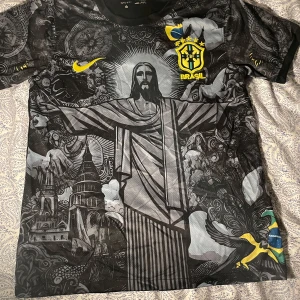 Svart Brasilien fotbollströja med mönster - Unik svart fotbollströja från Nike med Brasilien-tema. Tröjan har ett detaljerat mönster med Kristusstatyn och andra ikoniska motiv. Den har korta ärmar och det brasilianska landslagets emblem i gult och grönt. Perfekt för fotbollsfans som vill sticka ut!