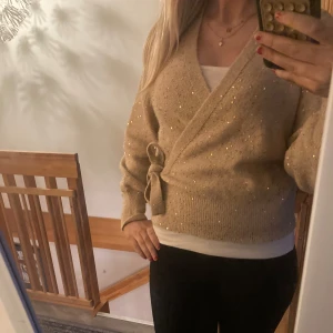 Beige omlottkofta med glitterdetaljer - Säljer en snygg beige omlottkofta med glittriga detaljer. Koftan har långa ärmar och ett knytband i midjan för en justerbar passform. Perfekt för att lägga till lite glamour i vardagen. ✨