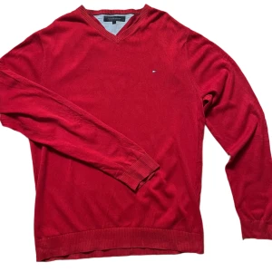 Röd tröja från Tommy Hilfiger - Snygg röd tröja från Tommy Hilfiger med v-ringning och diskret logga på bröstet. Tröjan har långa ärmar och ribbade muddar vid ärmslut och nederkant. Perfekt för en stilren look.