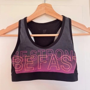 Svart sport-bh med rosa text - Säljer en svart sport-bh från H&M med rosa text 'BE STRONG BE FAST' framtill. Den har en racerback-design och är tillverkad i ett stretchigt material som ger bra stöd under träning.