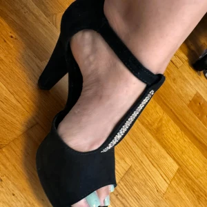 Svarta klackskor med strassdetaljer - Säljer ett par snygga svarta klackskor med öppen tå och strassdetaljer på remmen. Skorna har en bekväm passform och är perfekta för festliga tillfällen. De är i bra skick och redo att användas.