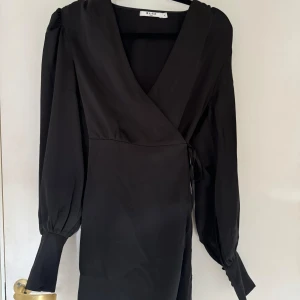 Svart omlottklänningen i satin  - Svart omlottklänningen i satin från Na-kd. I storlek 36. Använd 1 gång. Slutsåld på hemsidan.