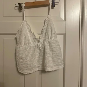 Vit ärmlös topp med knytning - Säljer en söt vit ärmlös topp med smala axelband och knytning framtill. Toppen har en lätt textur och är perfekt för sommaren. Den är i bra skick och passar både till vardags och fest.
