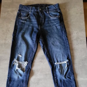 Blå jeans från G-Star RAW - Säljer ett par snygga blå jeans från G-Star RAW. De har en cool slitning på knäna och en klassisk femficksdesign. Jeansen är i en straight fit och har en knappgylf. Perfekta för en avslappnad stil!