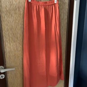 Orange långkjol - Säljer en superfin orange långkjol. Den är enkel och stilren med en elastisk midja som gör den bekväm att bära. Perfekt för sommaren eller våren! Kjolen är i ett lätt och luftigt material, vilket gör den idealisk för varma dagar. Passar både till vardags och fest. Den har slitsar på båda sidorna och fickor. 