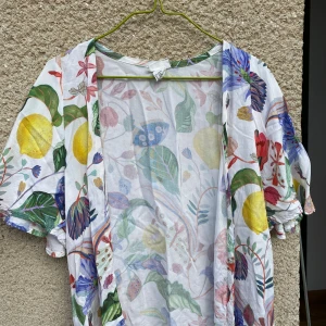 Färgglad blommig kimono - Säljer en superfin och färgglad kimono med blommigt mönster. Den är perfekt för sommaren med sina korta ärmar och lösa passform. Materialet känns lätt och luftigt, vilket gör den idealisk för varma dagar. Den har en öppen design utan knappar eller dragkedja.