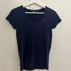 Mörkblå t-shirt från Ralph Lauren - Säljer en snygg mörkblå t-shirt från Ralph Lauren med en klassisk v-ringning. Den är i storlek xs. Den har en liten röd logga på bröstet som ger en stilren touch. Perfekt för både vardag och fest!