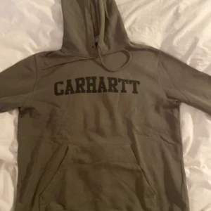 Grön hoodie från Carhartt - Säljer en snygg grön hoodie från Carhartt i storlek M. Den har en klassisk design med Carhartt-loggan tryckt över bröstet. Hoodien är perfekt för höst och vinter med sin mysiga och varma känsla. Den har en stor ficka framtill och en justerbar huva. Perfekt för vardagsbruk eller chilliga dagar!
