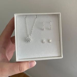 Smyckeset med halsband och örhängen i silver - Säljer ett elegant smyckeset som inkluderar ett halsband och två par örhängen i silver. Halsbandet har en vacker snöflingeformad hängsmycke och örhängena matchar perfekt med små snöflingor och pärlor. Perfekt för både vardag och fest!