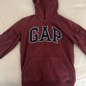 Vinröd hoodie från GAP - Säljer en snygg vinröd hoodie från GAP i storlek 14-16. Den är gjord av 77% bomull och 23% polyester, varav 9% är återvunnen polyester. Hoodien har en dragkedja framtill och en stor GAP-logga i vitt och svart på bröstet. Perfekt för höst och vinter! 🧥