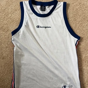 Vit linne från Champion - Säljer en vit linne från Champion med marinblå detaljer. Linnet har ett sportigt utseende med meshmaterial och Champion-loggan på bröstet. Perfekt för träning eller en avslappnad dag.