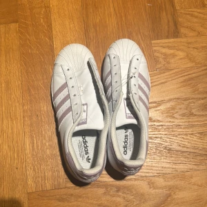 Vita Adidas Superstar slip-on - Säljer ett par vita Adidas Superstar slip-on med lila ränder. Skorna har den klassiska snäckformade tån och är tillverkade i syntetmaterial. Perfekta för en stilren och bekväm look. Aldrig använda men snören finns inte❤️