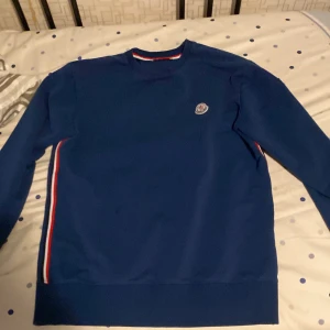 Blå tröja med ränder från Moncler - Säljer en snygg blå tröja från Moncler med ränder i rött och vitt längs sidorna. Tröjan har långa ärmar och ett Moncler-märke på bröstet. Perfekt för en stilren look.