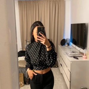 Svart prickig croppad blus - Säljer en snygg svart blus med vita prickar. Blusen är croppad och har långa ärmar med resår vid ärmsluten. Den har en knytning i ryggen som ger en fin detalj. Perfekt för en trendig look!